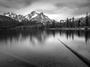 Black and white landscape of the Sawtooth Mountains reflecting in the still waters of a forest lake in Idaho, with dramatic clouds above and fallen logs in the foreground