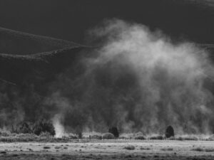 Black and white landscape photograph of morning mist rising from a valley floor in Idaho, with dramatic light illuminating low-lying fog against the backdrop of dark, rolling hills and scattered trees.
