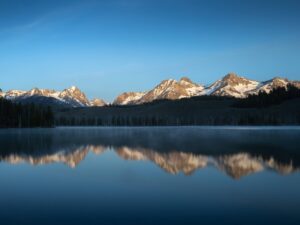 Peaceful alpine sunrise at an Idaho mountain lake, with snow-covered Sawtooth peaks bathed in golden light and reflected in the still, misty water under a clear blue sky.