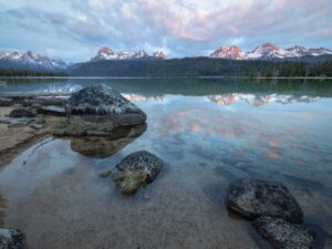 Tranquil sunrise over Redfish Lake in Idaho, with the Sawtooth Mountains reflected in the crystal-clear water. Foreground features granite boulders and sandy shoreline, with soft pastel clouds illuminating the alpine peaks.