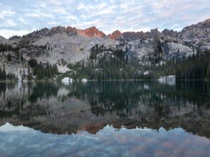 Alice Lake in Idaho’s Sawtooth Mountains at sunrise, with jagged granite peaks glowing pink above a calm alpine lake reflecting pine trees and soft morning clouds.